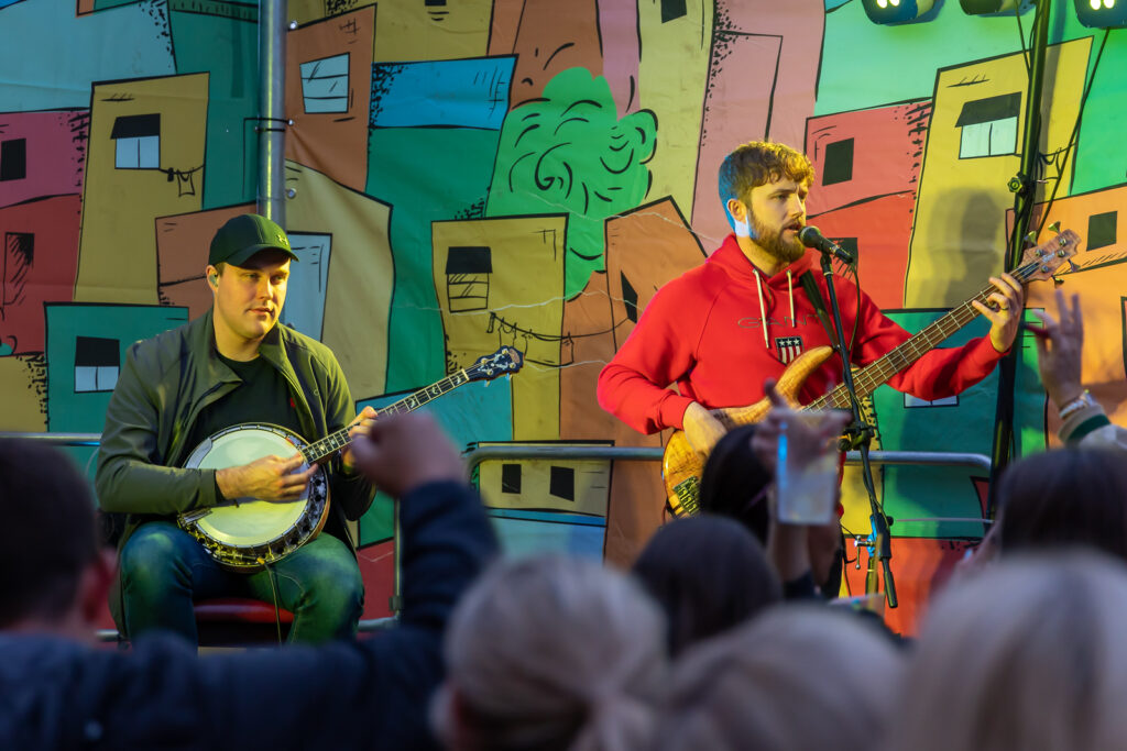 Two male musicians performing on stage. One man sitting down playing the banjo and other man is standing and playing bass guitar. Colourful backdrop to the stage. Musicians looking somber. Backs of people's heads in the crowd looking at the musicians performing on stage.