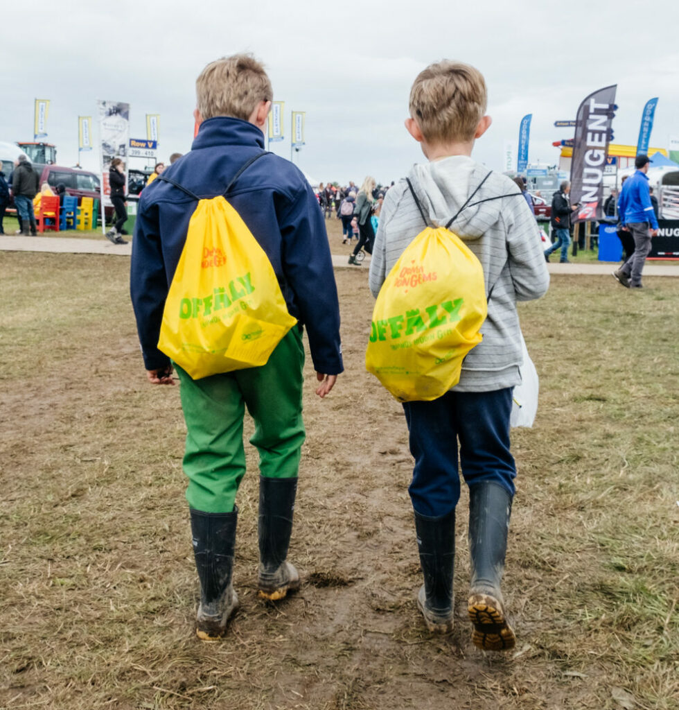 Two young boys walking away from the camera. Each wearing muddy wellies and wearing an Offaly Hidden Gem bright yellow drawstring bag on their backs.
