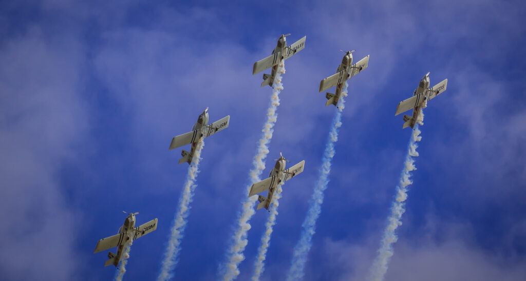 6 aircraft flying through a bright blue sky. A line of smoke trailing behind each aircraft.