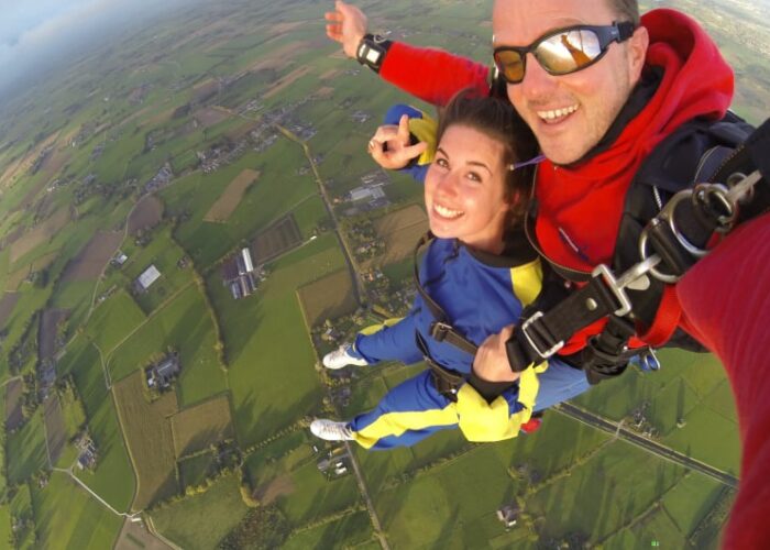 Man And Woman Skydiving Above Green Fields