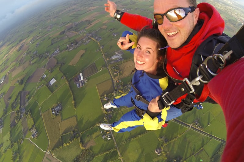 Man And Woman Skydiving Above Green Fields