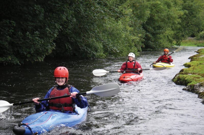 Three People Kayaking Down A Wooded River