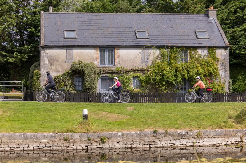 Three people cycling past an ivy covered stone cottage