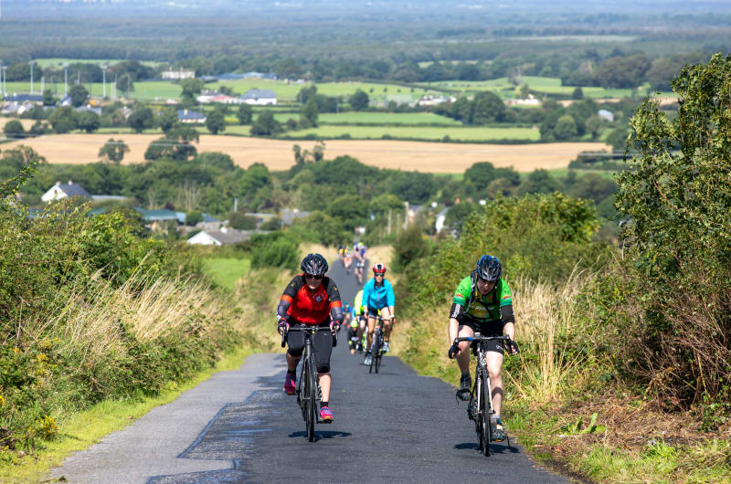 Cyclists Climbing A Rural Road Through Green Fields lhnlfz