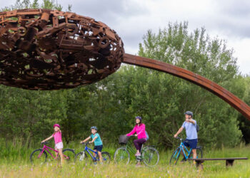 Family Cycling Under A Large Rusty Metal Sculpture
