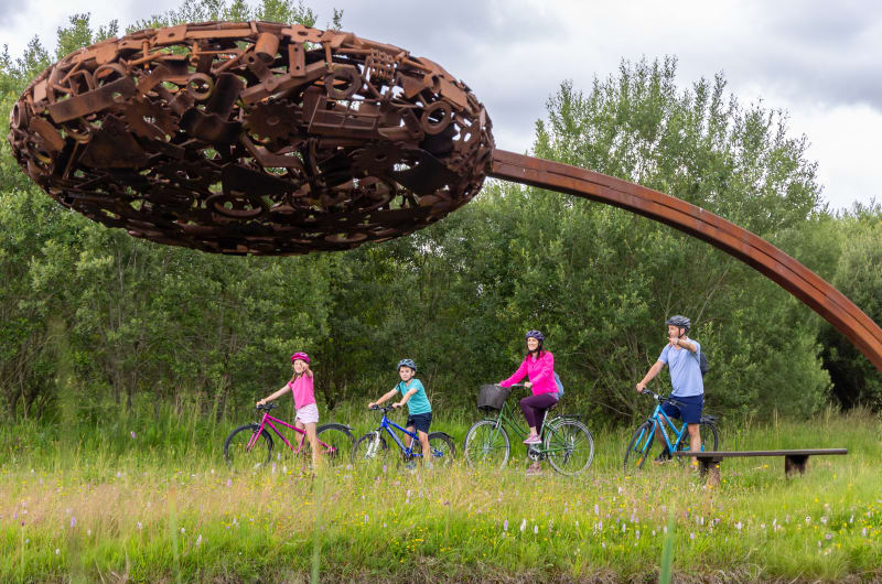Family cycling under a giant rusted metal sculpture w4lphx