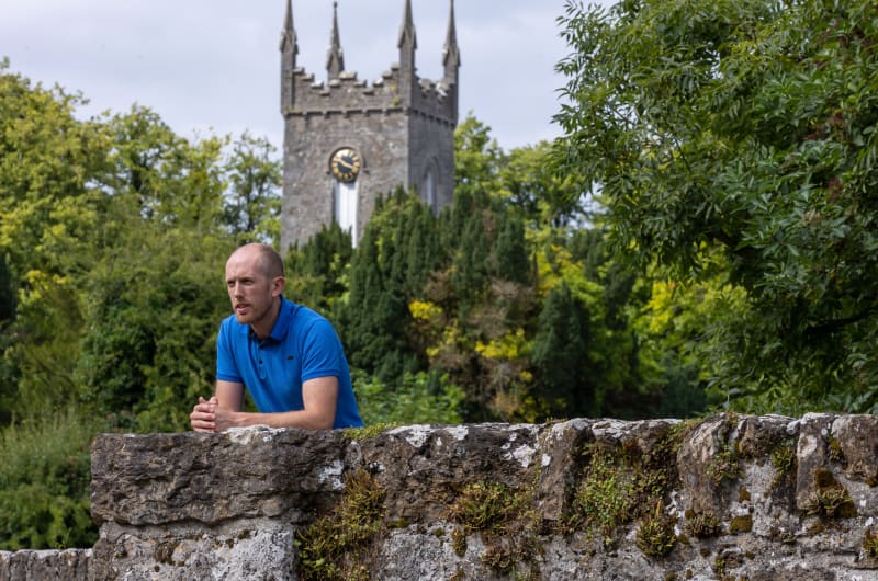 Man in blue shirt leaning on stone wall tf8wxo
