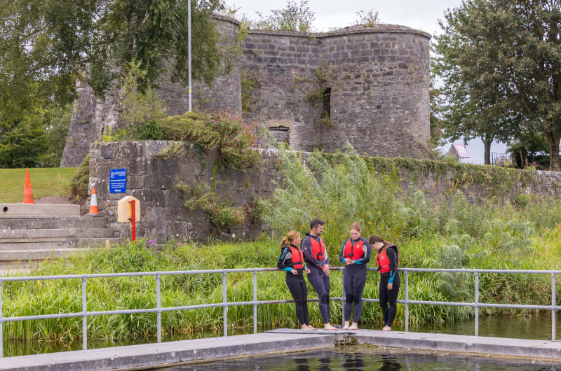 People in wetsuits near historic stone tower pm4a38