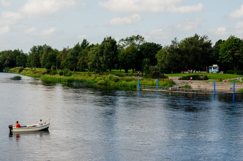 Small Boat On River With Grassy Shoreline Trees fwrr0q