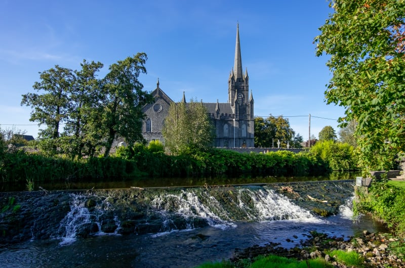 Stone cathedral spire behind flowing river weir greenery mbepit