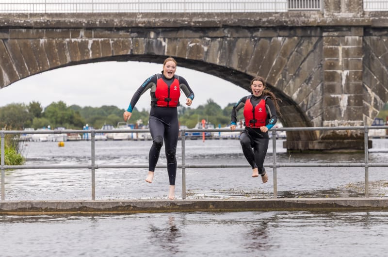 Two Girls In Wetsuits Jumping From Bridge Ledge sqjl4i