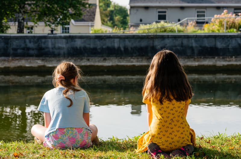 Two Girls Sitting On Grass Facing A Canal k2f8ul