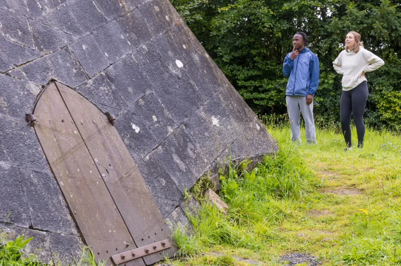 Two people standing near slanted stone structure door fvb8eo