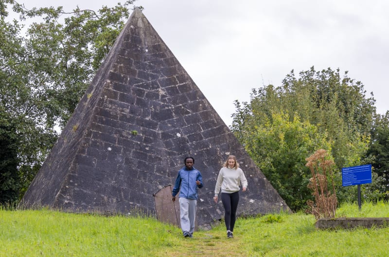 Two people walking in front of stone pyramid kcq68q