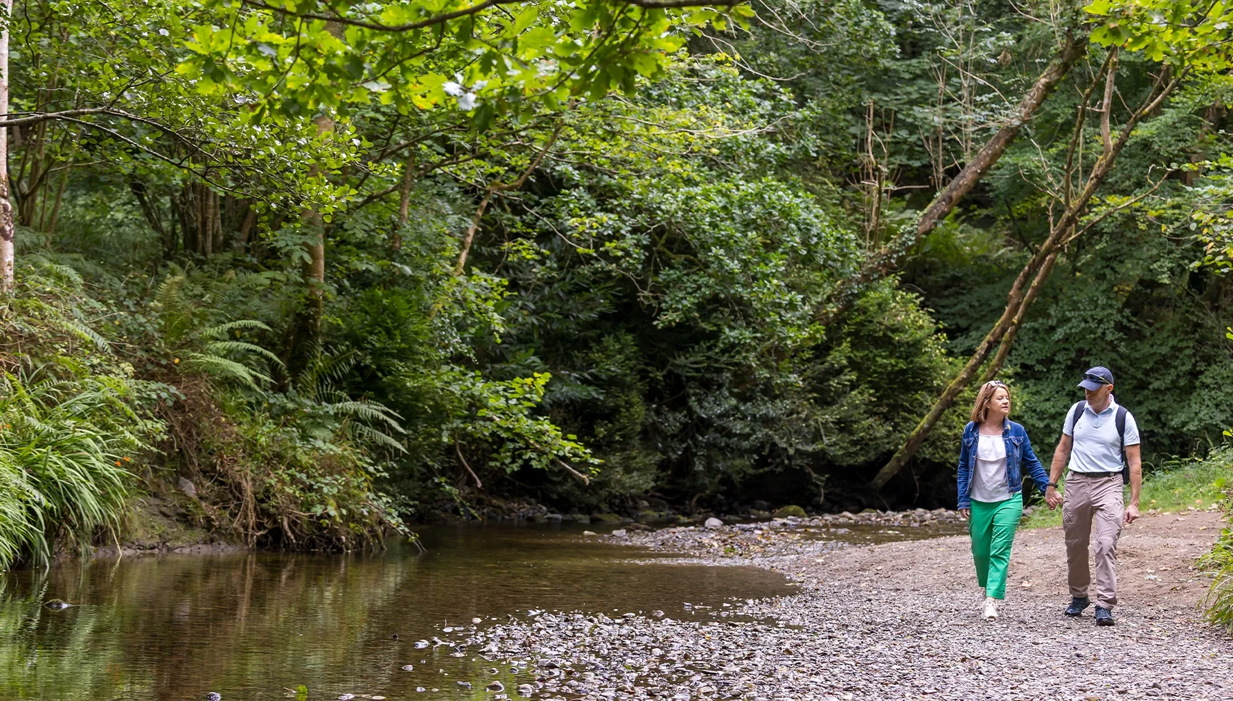 Man and woman walking holding hands on stony riverside in forest